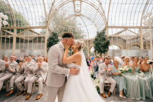 Couple share first kiss surrounded by guests in the glass Orangery at Tortworth Court wedding